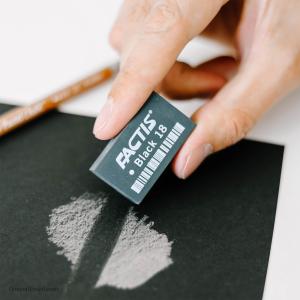 A hand uses a Factis® Magic Black™ Eraser to remove white pencil marks from black paper, with a brown pencil resting nearby.