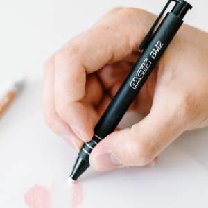 A hand is holding a black mechanical eraser labeled "FACTIS" with the model number "BM-2." The hand appears to be erasing or shading a drawing a pink swatch on white paper. Next to the hand lies a brown wooden pencil branded "General's® MultiPastel®."