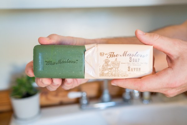 A person holds a partially unwrapped green bar of "The Masters" soap in front of a wooden shelf with small plants in the background. The soap wrapper displays vintage-style text and artwork.