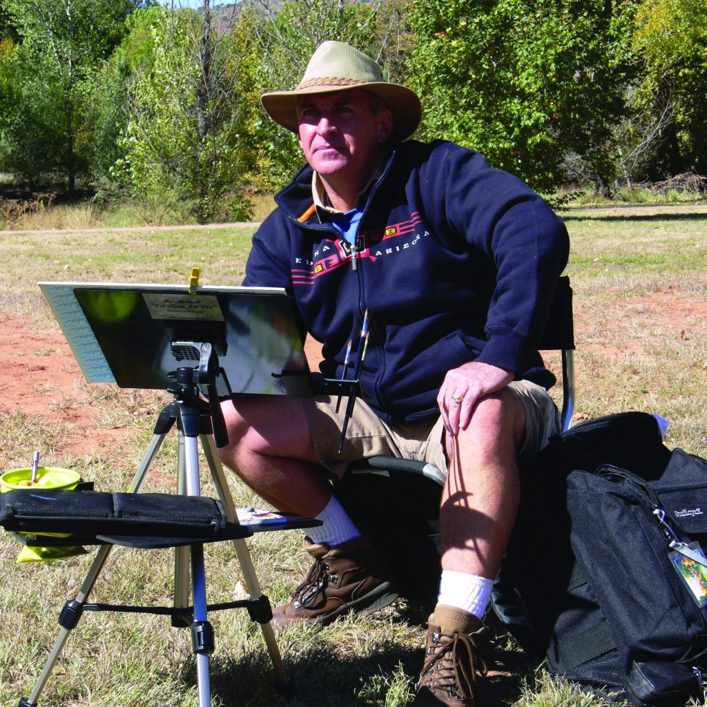 Artist Tom Lynch wearing a hat and a dark jacket sits on a portable chair outdoors, painting on a canvas attached to an easel. The scene is set in a grassy area with trees in the background. The person is surrounded by painting supplies, including a backpack and brushes.