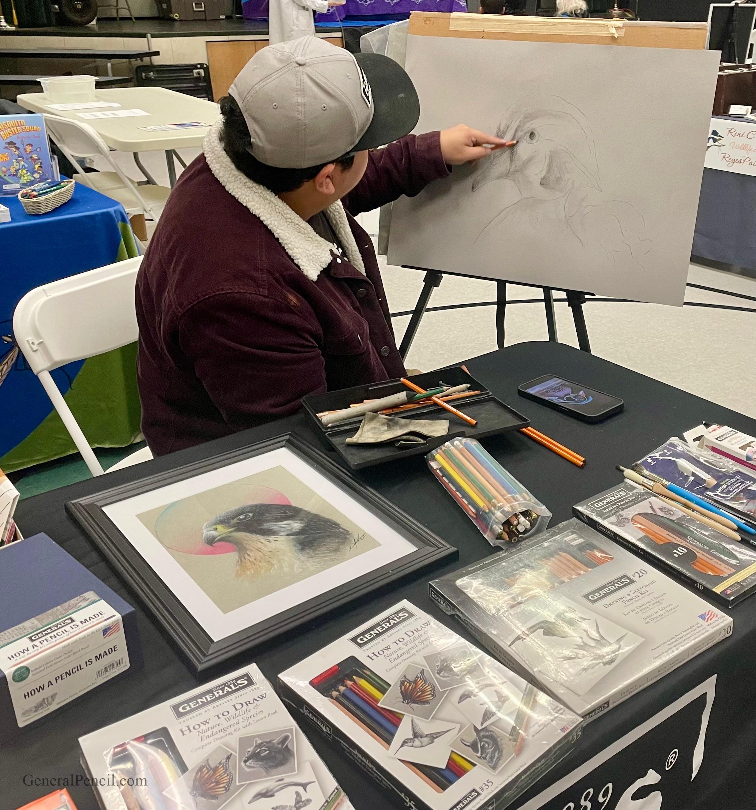 A person wearing a cap and jacket sketches a bird on a large drawing pad at a table covered with colored pencils, drawing supplies, and a framed bird illustration at an art event.