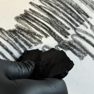 A gloved hand holds a piece of charcoal, drawing bold black strokes on white paper. Several overlapping, textured charcoal lines are visible in the background.