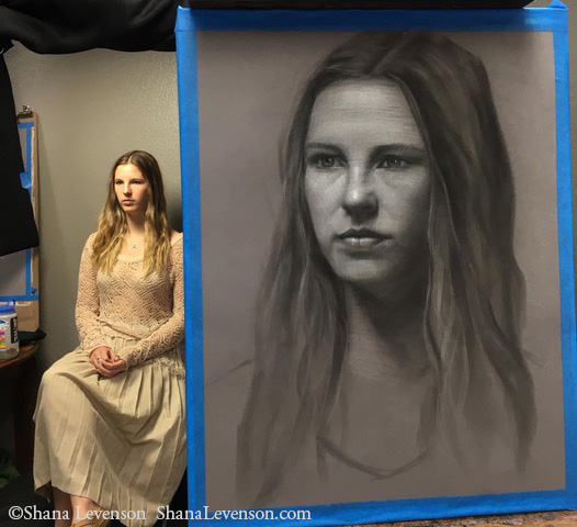 A woman with long, wavy hair and a light-colored dress sits beside a large, detailed pencil portrait of herself, which is taped to an easel. The portrait closely resembles her serious expression.