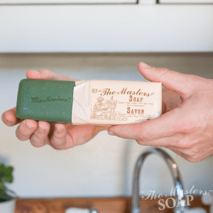 A person holds a green bar of "The Master's Soap," partially unwrapped from its beige paper packaging, standing in a bright kitchen with a sink and small plant in the background.