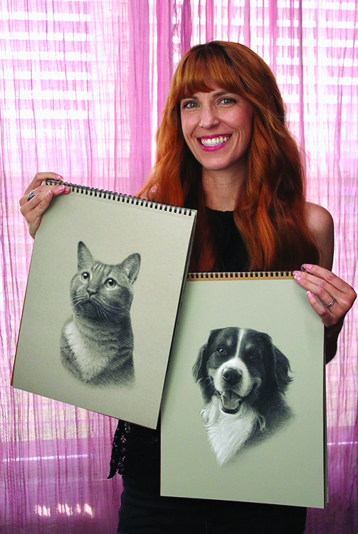 A woman with long red hair smiles while holding two sketchpads, one with a pencil drawing of a cat and the other with a pencil drawing of a dog, in front of pink curtains.